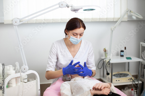Young female doctor cosmetologist in protective mask and blue rubber gloves makes facial massage and skin treatment for young woman client. Spa face massage in beauty salon  facial beauty treatment