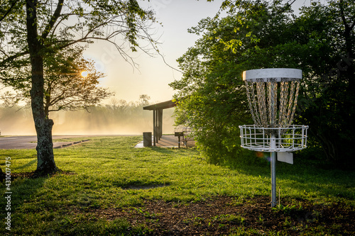 Disc golf target in a veterans park in Lexington, Kentucky during early morning hours. Sun is shining through the fog on the background.