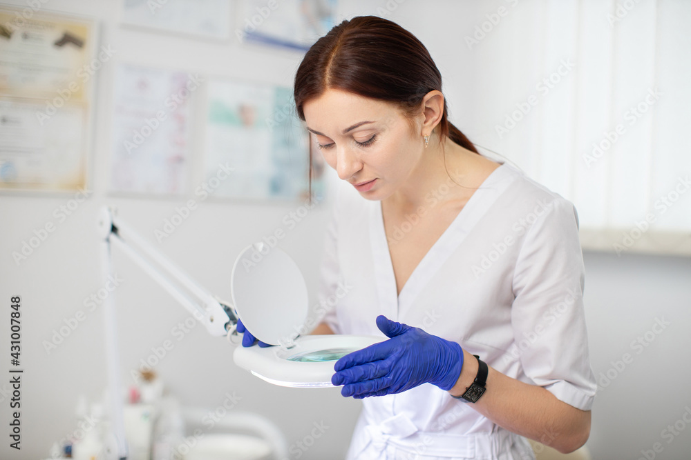 Professional skin problems. Close up portrait of pretty confident female cosmetologist in white uniform and blue protective gloves, looking through magnifying lamp, working at modern clinic Professional skin problems. Close up portrait of pretty confident female cosmetologist in white uniform and blue protective gloves, looking through magnifying lamp, working at modern clinic