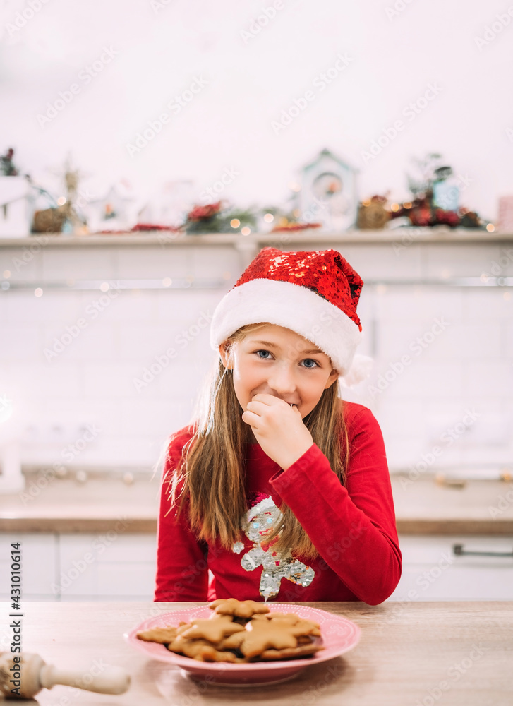 Vertical portrait of a girl who eats gingerbread cookies in the kitchen in a red sweater and a Santa hat. Place for the text. Vertical portrait of a girl who eats gingerbread cookies in the kitchen in a red sweater and a Santa hat. Place for the text.