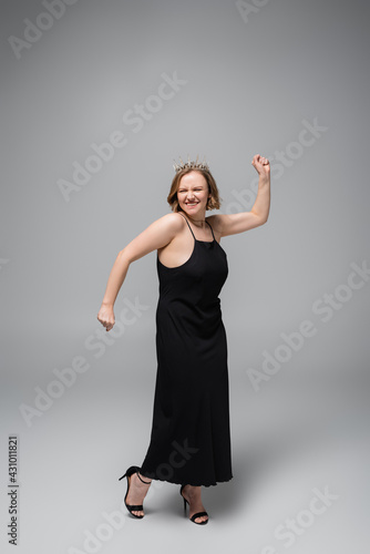 full length of happy plus size woman in black slip dress and crown on grey