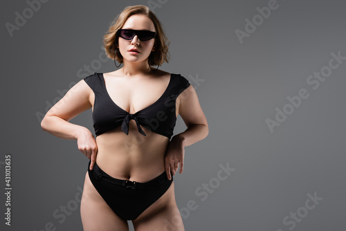 overweight woman in sunglasses and black swimsuit posing isolated on grey © LIGHTFIELD STUDIOS