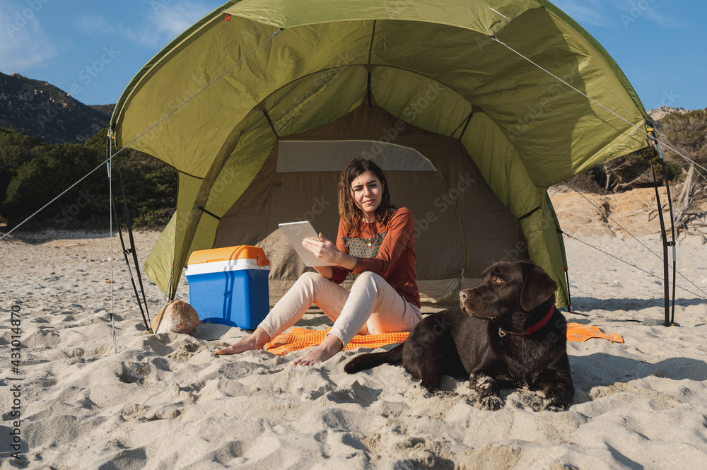 Young attractive woman in a camping beach with tent sitting on the sand holding a tablet and looking at the dog lying next to her.