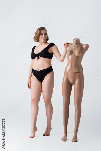 full length of overweight and barefoot young woman smiling near plastic mannequin on white