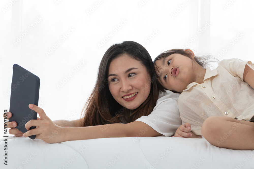 Fototapeta premium Young little girl playing with her mother on bed, she is lying on her mother back looking at tablet screen. Young mother and daughter talking on bed while playing with computer tablet.
