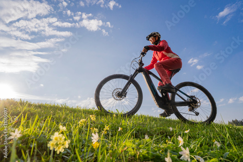 pretty mid age woman riding her electric mountain bike in early springtime in the Allgau mountains near Oberstaufen  in warm evening light with blooming spring flowers in the Foreground