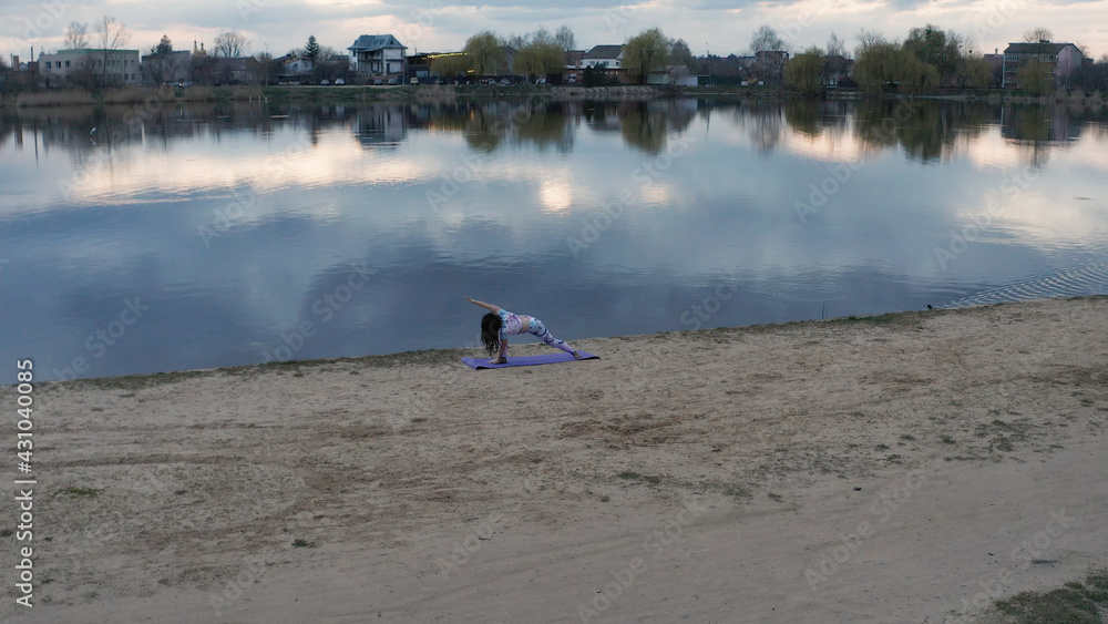 A young woman, yogina practicing yoga on the beach in the nature near the lake at sunset. 