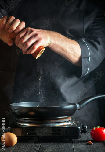 Chef or cook adds pepper while cooking eggs in a pan. Work environment on vintage kitchen table. Vertical image