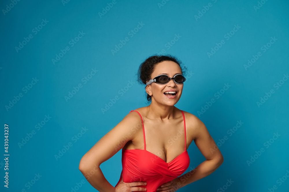 Young woman swimmer in red bathing suit and swimming goggles smiling toothy smile on blue background Young woman swimmer in red bathing suit and swimming goggles smiling toothy smile on blue background