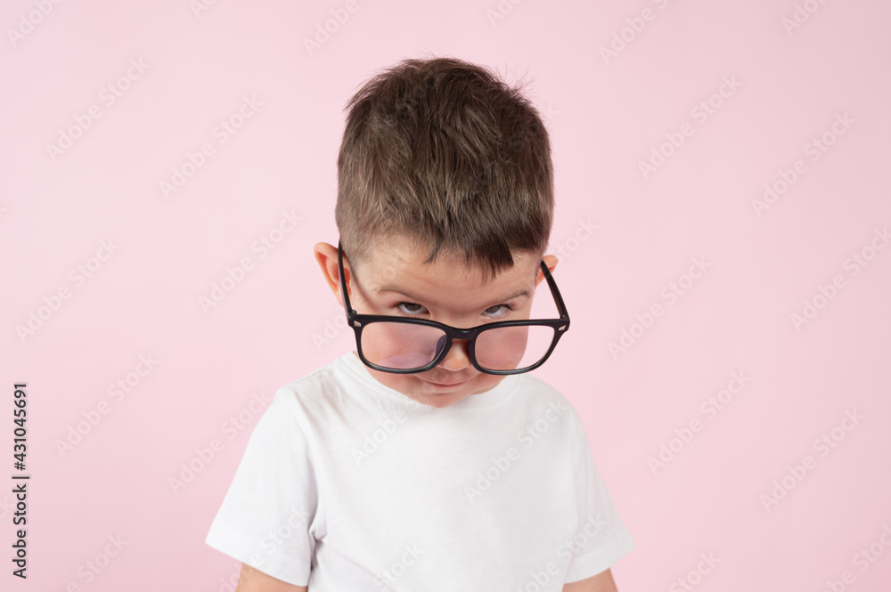 A serious little boy peeks out from under his glasses, close up portrait on pink background.
