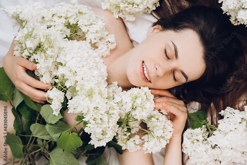 Close up portrait of charming beautiful young woman with closed eyes and sincere smile lying in comfy white bed, spring flowers around head. Relaxed and cute girl with clear skin enjoys, feels joy