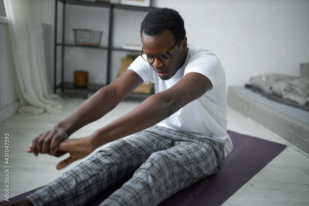 Portrait of lazy young black male with inflexible body sitting on mat in comfortable clothing doing forward bend, trying to touch feet with hands. Sleepy African guy practicing morning yoga Portrait of lazy young black male with inflexible body sitting on mat in comfortable clothing doing forward bend, trying to touch feet with hands. Sleepy African guy practicing morning yoga