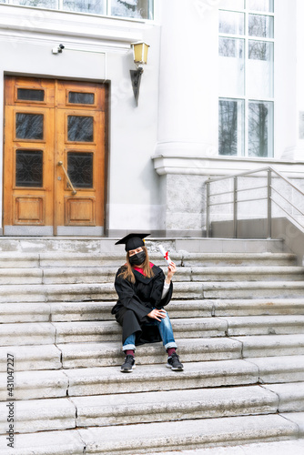 Young woman student in black graduation gown, graduation cap with yellow tassel, protective face mask sitting on steps of educational institution and holding roll of certificate with red ribbon,