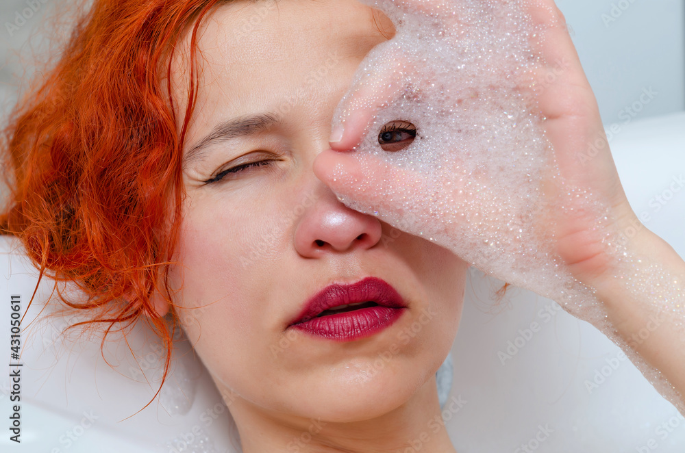 Fototapeta premium Portrait of a young beautiful woman in bathroom, looking through foam between fingers