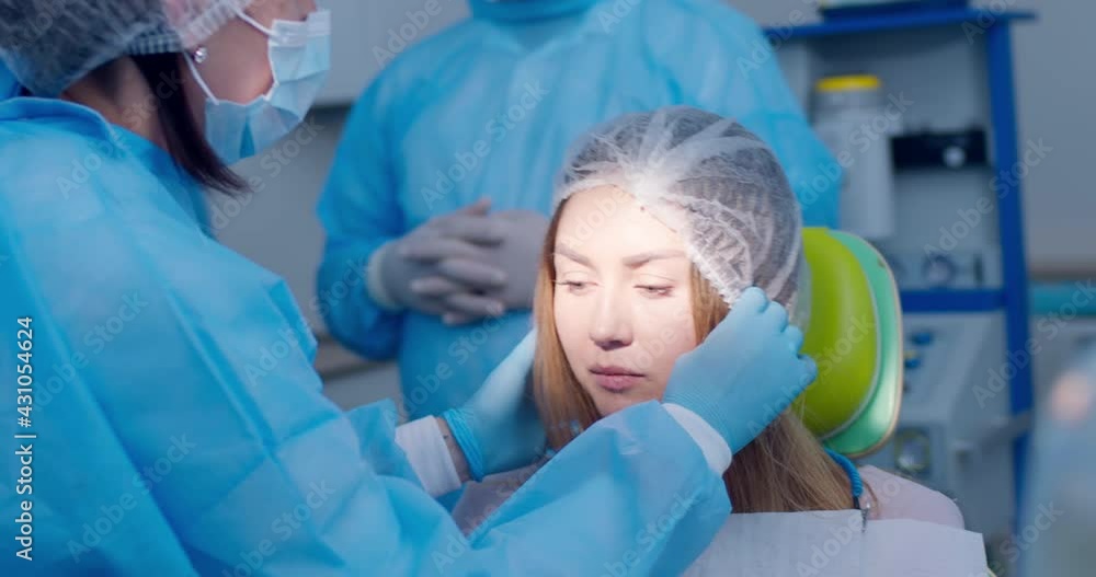 A female patient is sitting in the dental chair with a disposable napkin while a female medical worker is putting on her head a disposable hat. A female patient is sitting in the dental chair with a disposable napkin while a female medical worker is putting on her head a disposable hat.