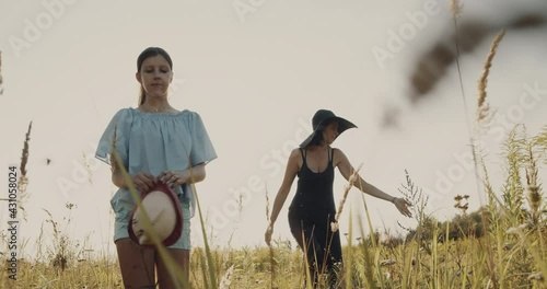 mother and daughter, walk on a yellow summer lawn under the sun. Girls going straight ahead slowly. Holding hat in a hand. Nature background, weekend out of the city