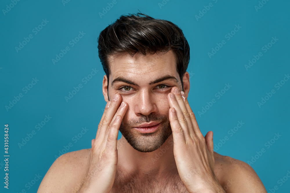 Beauty portrait of handsome man looking at camera with a smile while applying moisturizing cream on his face isolated over blue background Beauty portrait of handsome man looking at camera with a smile while applying moisturizing cream on his face isolated over blue background