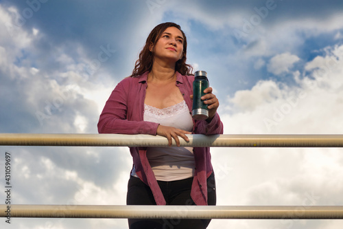 Hermosa mujer colombiana pensativa en un puente con un termo con agua en la mano en el atardecer