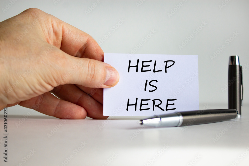 Help and support symbol. White paper with words 'Help is here' in businessman hand, metalic pen. Beautiful white background. Business, help and support concept. Copy space. Help and support symbol. White paper with words 'Help is here' in businessman hand, metalic pen. Beautiful white background. Business, help and support concept. Copy space.