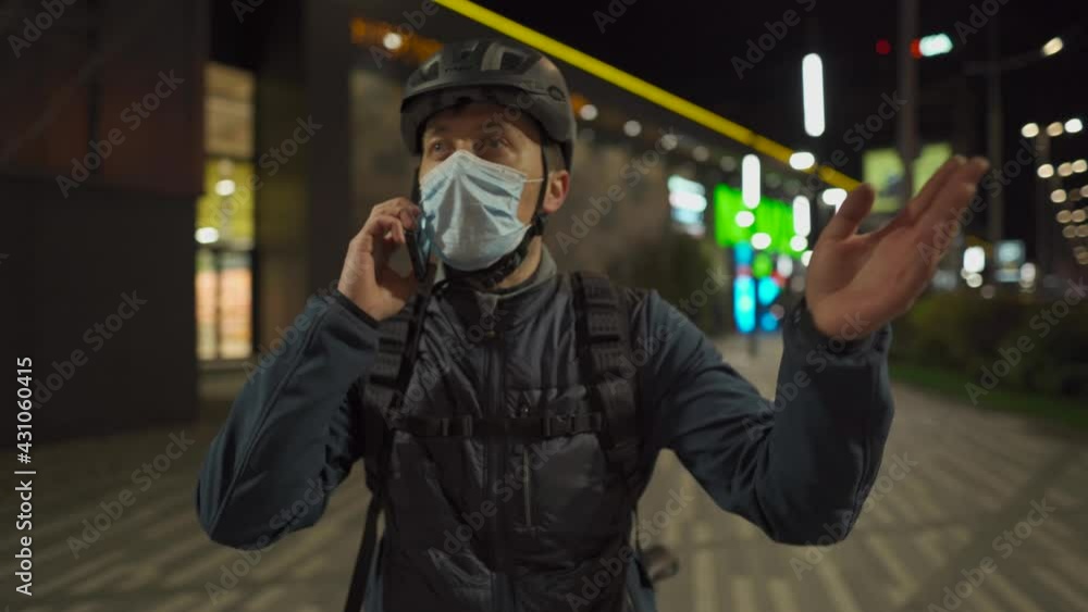 Cyclist in helmet and protective mask commuter with bicycle on way home from work talking on phone at night city. Deliveryman on bike, delivery parcels, make phone call during coronavirus quarantine Cyclist in helmet and protective mask commuter with bicycle on way home from work talking on phone at night city. Deliveryman on bike, delivery parcels, make phone call during coronavirus quarantine
