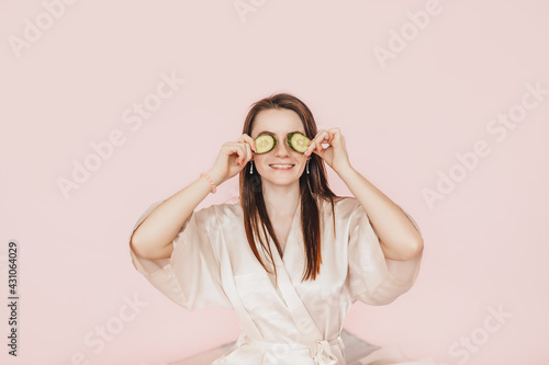 Girl make homemade face beauty masks. Cucumbers for the freshness of the skin around the eyes. Woman take care of youthful skin. Model laughing and having fun in spa on pink background