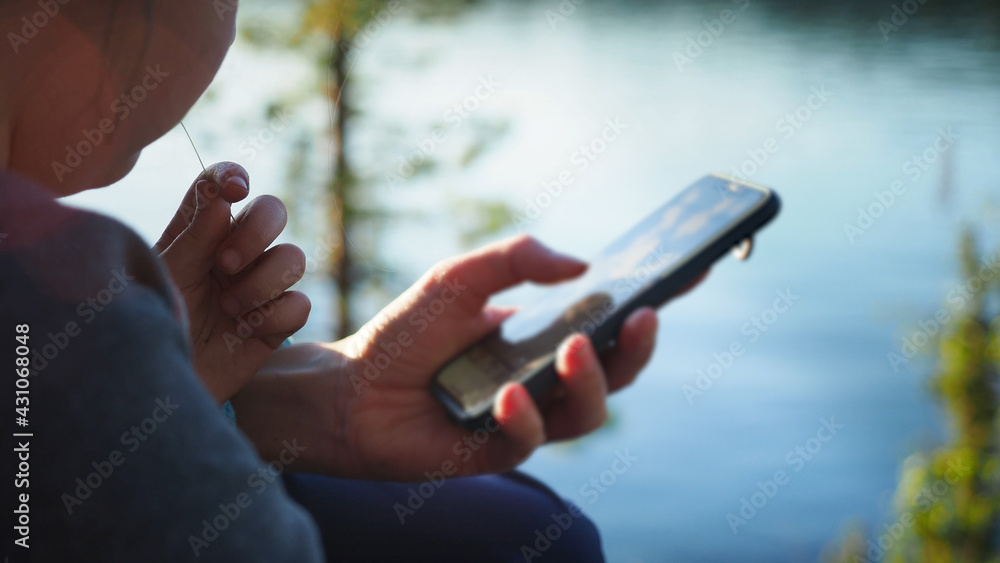 Close up. Mother using her mobile phone apps browsing chatting playing web surfing network with little daughter opposite the lake during sunset on spring camping vacation. Concept of modern technology
