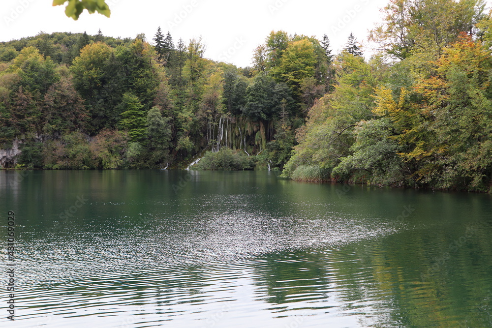 Waterfall with powerful streams against the backdrop of a lake and a mountain slope overgrown with moss, trees with autumn foliage are reflected in calm emerald water, Plitvice Lakes National Park Waterfall with powerful streams against the backdrop of a lake and a mountain slope overgrown with moss, trees with autumn foliage are reflected in calm emerald water, Plitvice Lakes National Park