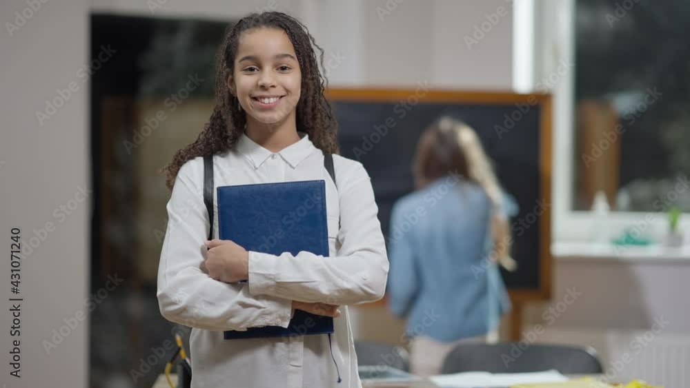 Middle shot portrait of positive cute African American schoolgirl with workbook posing in classroom with blurred Caucasian classmate at background. Happy confident girl standing in school indoors Middle shot portrait of positive cute African American schoolgirl with workbook posing in classroom with blurred Caucasian classmate at background. Happy confident girl standing in school indoors