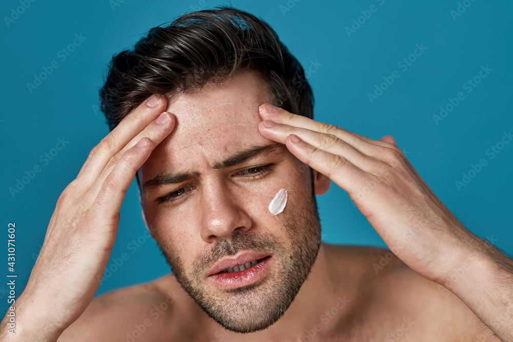 Beauty portrait of serious confident guy with brown hair looking aside while applying cream on his face, posing isolated over blue background