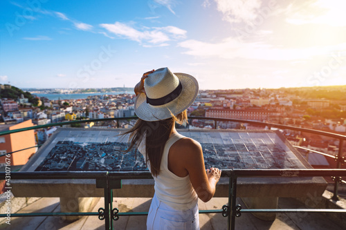 Young carefree woman tourist in white hat looking at Lisbon city from viewpoint at sunset. Tourism in Europe. Travel, vacation and holidays concept. 