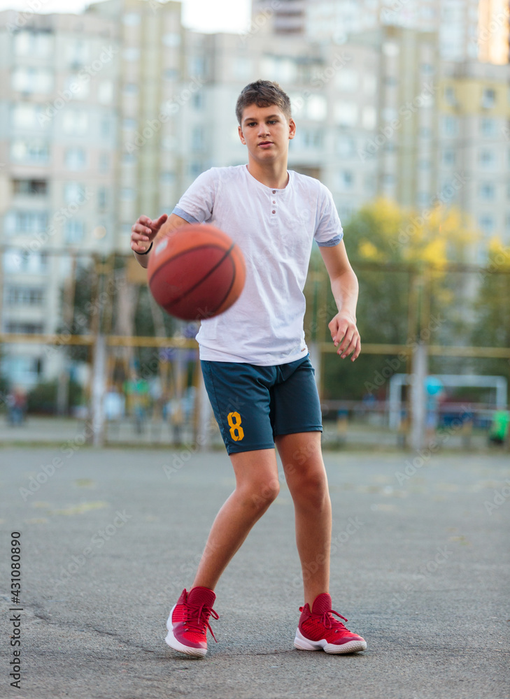 A cute young boy plays basketball on the street playground in summer. Teenager in a white t-shirt with orange basketball ball outside. Hobby, active lifestyle, sports activity for kids.