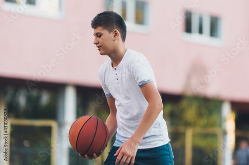 A cute young boy plays basketball on the street playground in summer. Teenager in a white t-shirt with orange basketball ball outside. Hobby, active lifestyle, sports activity for kids.