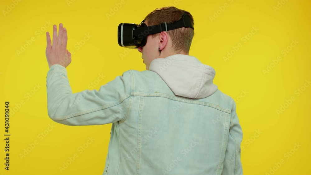 Rear view of teen stylish boy using virtual reality futuristic technology VR app headset helmet to play simulation 3D 360 video game, drawing. Young man on yellow studio background. People emotions Rear view of teen stylish boy using virtual reality futuristic technology VR app headset helmet to play simulation 3D 360 video game, drawing. Young man on yellow studio background. People emotions