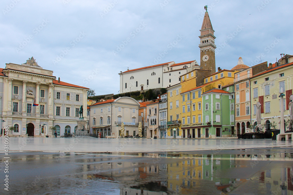 The main square of Piran with colorful houses, shops, cafes, a monument and a town hall with reflections in a puddle.