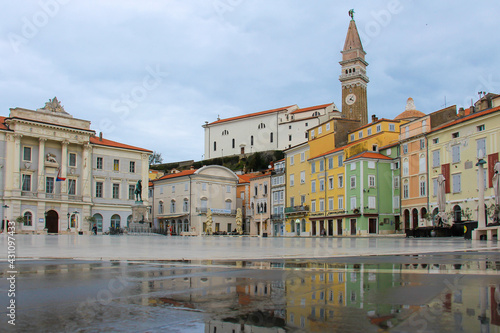 The main square of Piran with colorful houses, shops, cafes, a monument and a town hall with reflections in a puddle.