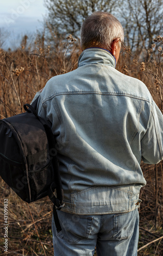 Elderly man in denim suit with black backpack looks for way through bushes while traveling through abandoned fields © Olga