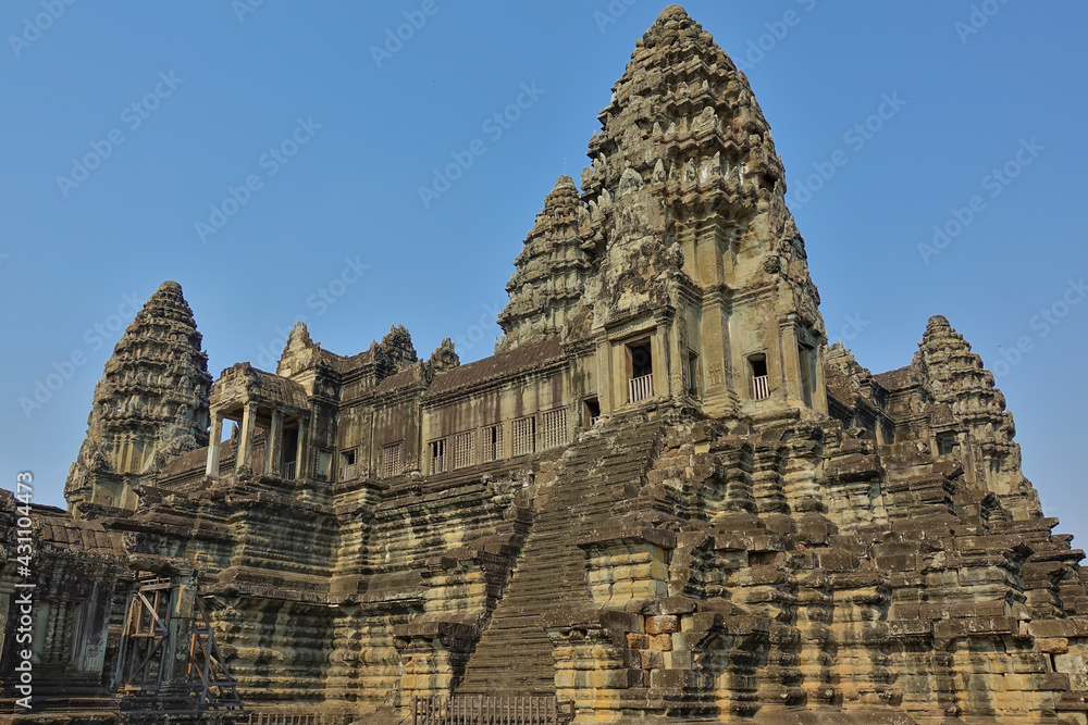 The ancient temple of Angkor against the blue sky. High towers are decorated with carvings and bas-reliefs. A stone staircase leads upstairs to a colonnaded terrace. Cambodia.
