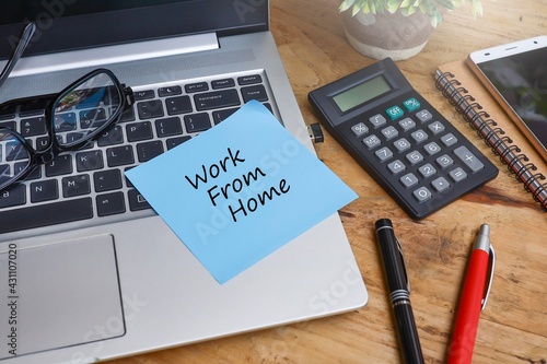 A piece of paper with a message on Work From Home, placed on the laptop keyboard with eyeglasses, a pen, calculator, and notebook on the wooden table. 