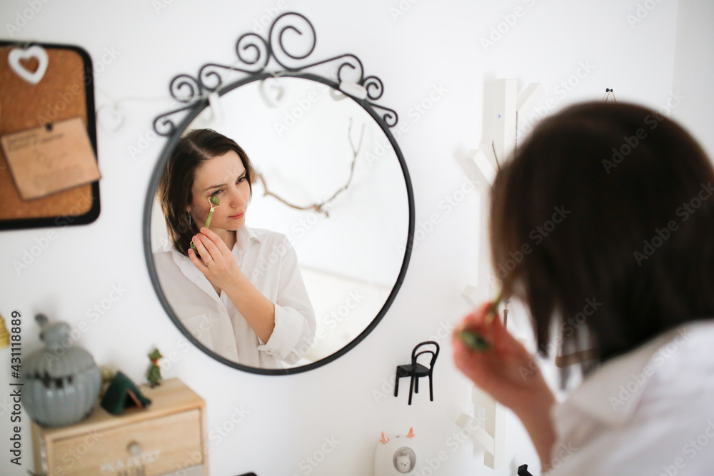 Cute caucasian woman with Jade Massage Roller near mirror ,female brunette does facial massage with roller, gouache. Concept beauty and self-care, skin and face care Cute caucasian woman with Jade Massage Roller near mirror ,female brunette does facial massage with roller, gouache. Concept beauty and self-care, skin and face care