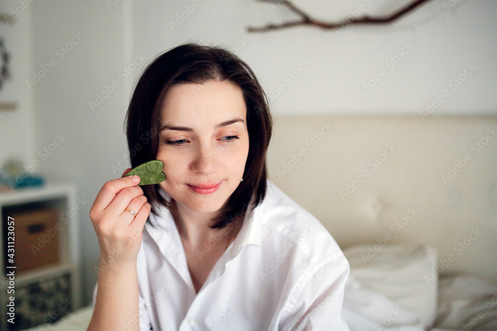 Cute caucasian woman with Jade gouache scraper in bedroom ,female brunette does facial massage with scraper gouache. Concept beauty and self-care, skin and face care Cute caucasian woman with Jade gouache scraper in bedroom ,female brunette does facial massage with scraper gouache. Concept beauty and self-care, skin and face care
