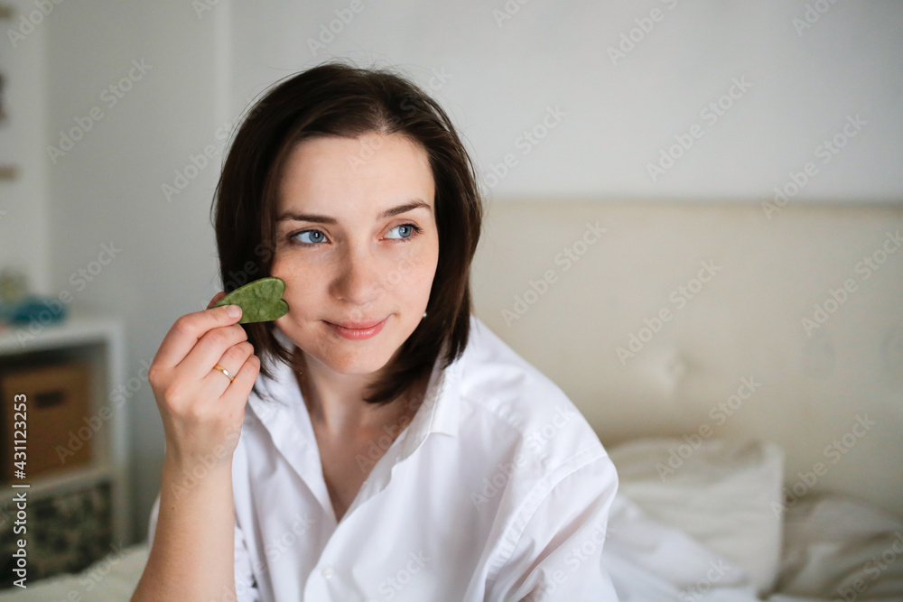 Cute caucasian woman with Jade gouache scraper in bedroom ,female brunette does facial massage with scraper gouache. Concept beauty and self-care, skin and face care Cute caucasian woman with Jade gouache scraper in bedroom ,female brunette does facial massage with scraper gouache. Concept beauty and self-care, skin and face care