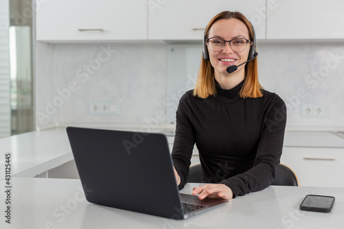A cheerful girl in a headset and headphones works in a call center behind a laptop, sitting at home in the kitchen. High quality photo