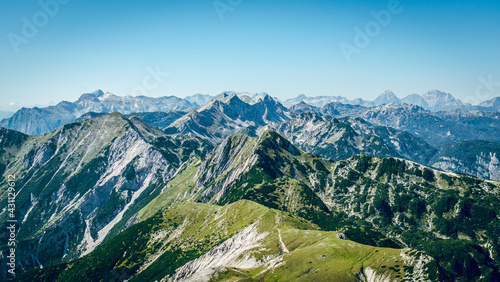 A panoramic view on the Alps from the top of mountain in Slovenia. Clear and sunny day. Sharp peaks around. A bit of haze in the valley. Outdoor activity. Lush green slopes with barren peaks. Calm