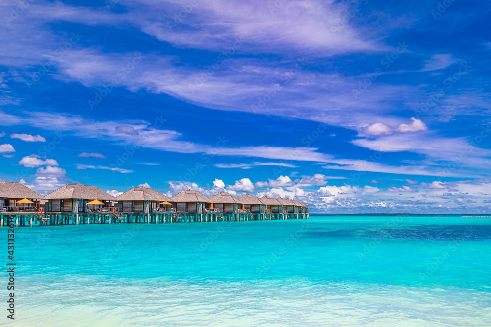 Island view of Maldives island, luxury water villas resort and wooden pier. Beautiful sky and ocean lagoon beach, palms sand. Summer vacation holiday and travel concept. Paradise shore coast landscape