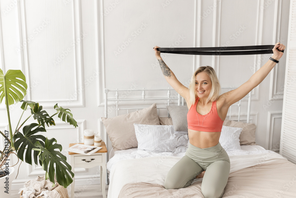 Happy young woman training in the morning, sitting on bed, doing exercises for the arms. Sportive blonde-hair girl stretching out, working out at home. Healthy lifestyle concept Happy young woman training in the morning, sitting on bed, doing exercises for the arms. Sportive blonde-hair girl stretching out, working out at home. Healthy lifestyle concept