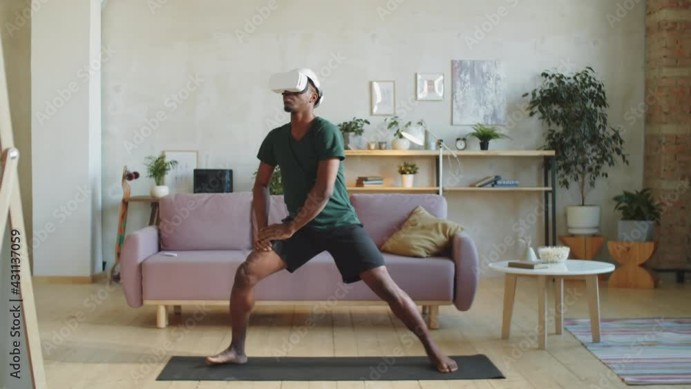 Young Afro-American man in VR headset and sportswear practicing warrior pose on mat at home while watching yoga workout in augmented reality Young Afro-American man in VR headset and sportswear practicing warrior pose on mat at home while watching yoga workout in augmented reality