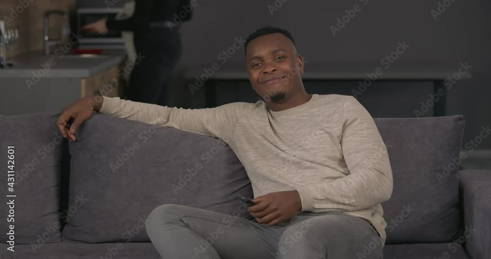 African man sitting on sofa and watching TV. Girlfriend wiping dishes on background. Cropped woman coming closer to husband and hits him with towel. Displeased man turnes off TV African man sitting on sofa and watching TV. Girlfriend wiping dishes on background. Cropped woman coming closer to husband and hits him with towel. Displeased man turnes off TV