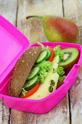 Whole grain roll with cheese, lettuce, tomato and cucumber and kiwi fruit and pear in pink lunch box
