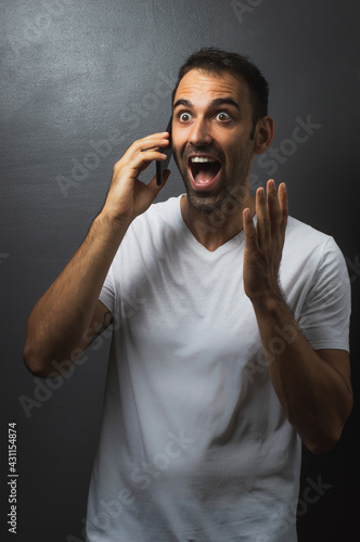 Man wearing a white tshirt over a grey background talking on the phone and looking surprised