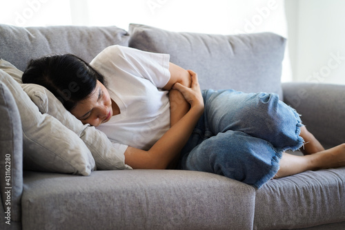 Young asian woman suffering from strong abdominal pain stomachache or period comes while lying on sofa at home